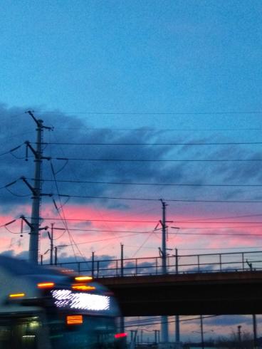 Color photograph. A blurred car drives past a metro track with electrical wires at sunset.
