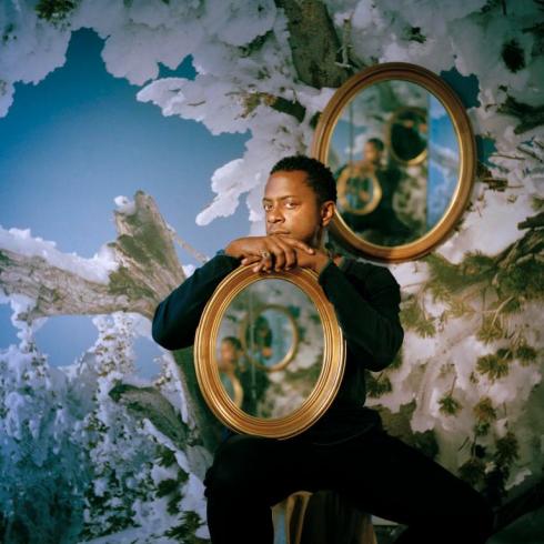Photo of Sanford Biggers sitting on a chair holding gold frame, with ornate floral background