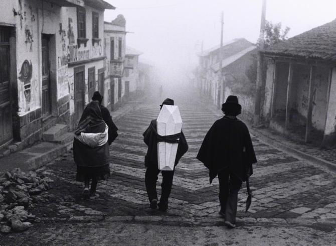 black and white photograph on cobblestone street of 3 people, with center figure holding small coffin