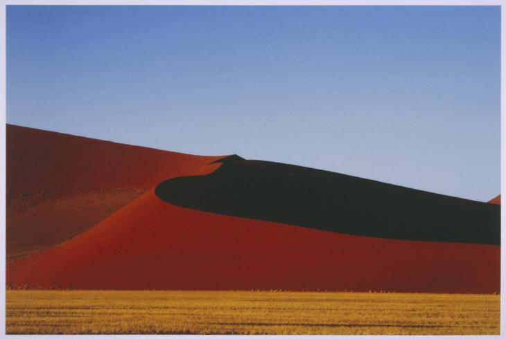minimalist photo of large red sand dune, yellow grown and blue sky