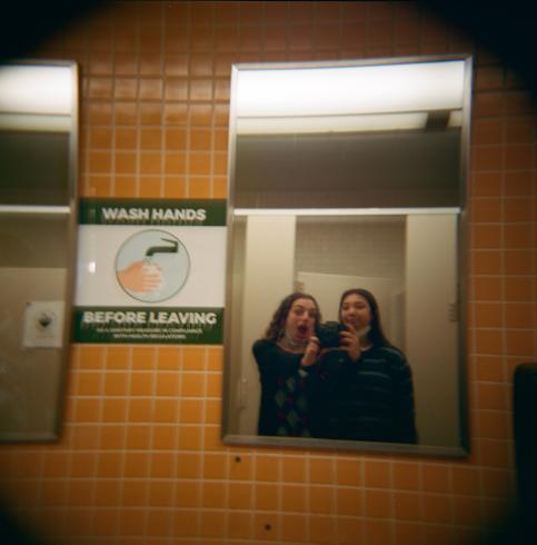 Image of two girls taking a bathroom selfie. The bathroom tile is mustard yellow