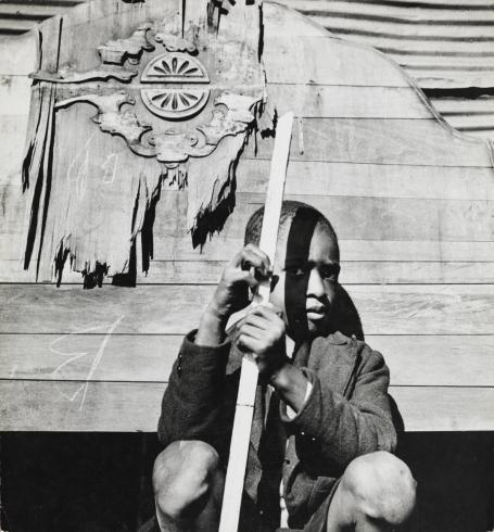 Black and white print of Black boy in a jacket holding a wooden stick in front of a wooden building