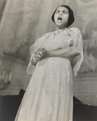 Black and white photograph of Marian Anderson holding her hands in white lacy dress while singing