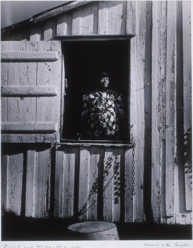 Black and white photograph of a wooden building with a Black woman in a floral dress wearing a hat