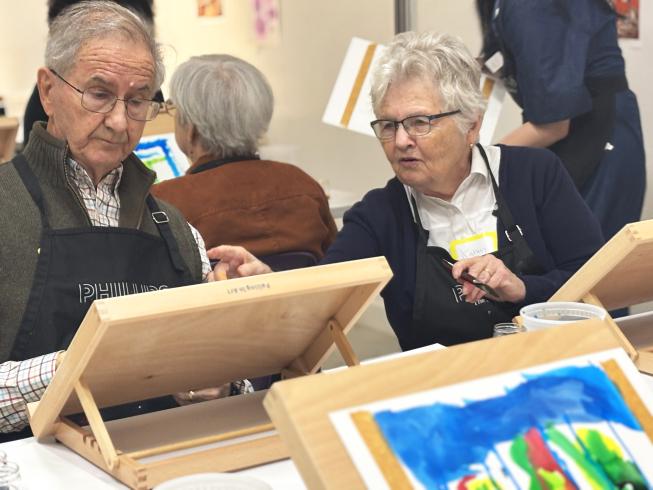 Two older adults sitting at a table and painting in an art studio